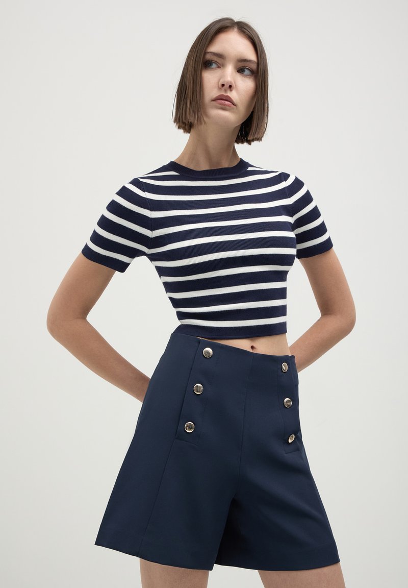 Giovane donna in posa con le mani sui fianchi, che indossa un crop top a righe blu e bianche con maniche corte e pantaloni corti blu con bottoni argento.