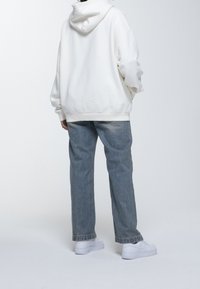 Sudadera blanca con capucha y un corte relajado, combinada con jeans de mezclilla azul claro y zapatillas blancas. Textura de tela suave visible.