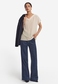 Beżowa bluzka krótki rękaw z dekoltem w serek, zestawiona z ciemnoniebieskimi szerokimi spodniami dżinsowymi. Model trzyma granatową kurtkę, stojąc na białym tle.