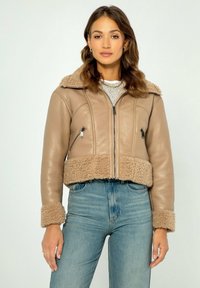 Giacca in pelle beige cropped con colletto e polsini in pelliccia, dotata di tasche con zip e una texture liscia. Indossata sopra jeans azzurri.