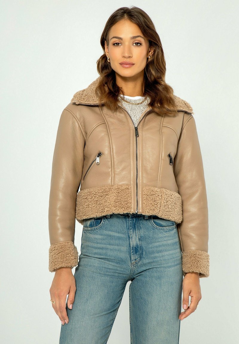 Giacca in pelle beige cropped con colletto e polsini in pelliccia, dotata di tasche con zip e una texture liscia. Indossata sopra jeans azzurri.