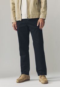 Beige lynlåsjakke over en hvid skjorte, kombineret med mørkeblå bukser og tanfarvede ruskindssko; glatte teksturer og bukser med lige ben.