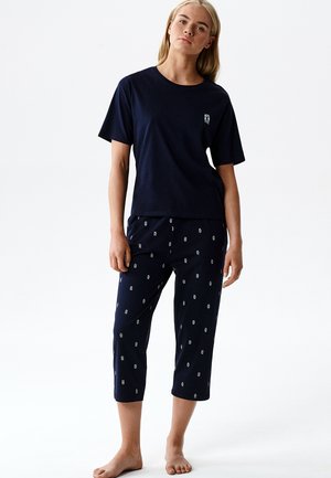 Kobieta stoi boso, ubrana w granatowy krótki rękaw i spodnie do połowy łydki z małymi białymi wzorami na jednolitym białym tle.