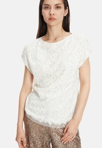 Weißes Spitzenshirt mit Blumenmustern und kurzen Ärmeln, kombiniert mit glitzernden, beige Paillettenhosen. Strukturiertes und geschichtetes Design.