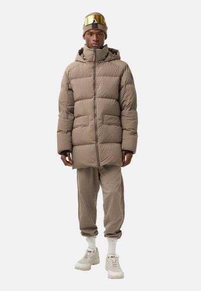 Beige puffer jacket with a high collar, full zipper, and two front pockets. Worn over matching beige sweatpants and white socks.
