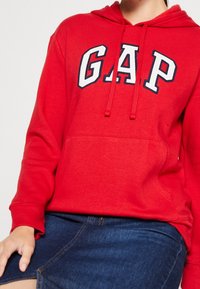 Sudadera roja con la palabra "GAP" en letras de bloque blancas y marinas. Confeccionada en tejido suave, cuenta con un bolsillo frontal tipo canguro y capucha con cordón.