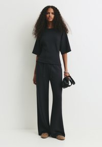 Femme aux longs cheveux bouclés portant un haut noir ample et un pantalon, tenant un petit sac à main noir, se tenant contre un mur blanc.