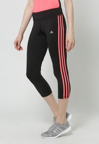 Person wearing black Adidas cropped leggings with pink side stripes and gray athletic shoes, standing against a plain background.