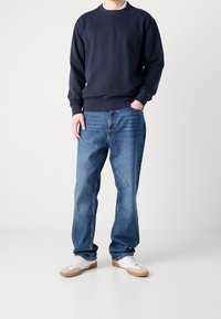 Námořnická mikina s žebrovanými manžetami v kombinaci se světle modrými džíny se širšími nohavicemi. Modelka má na sobě bílé tenisky s hnědými podrážkami. Jednoduchý design.