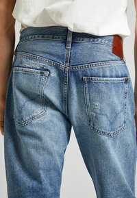Jeans de mezclilla azul con una textura desgastada, que presentan un parche de cuero marrón y dos bolsillos traseros con un mínimo desgaste.
