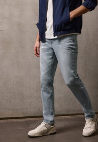 Man draagt lichtblauwe spijkerbroek, witte sneakers, wit T-shirt en een marineblauw jack, staand tegen een betonnen muur.