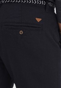 Black trousers made of textured fabric, featuring a back pocket with a brown button and a triangular leather patch with logo detail.