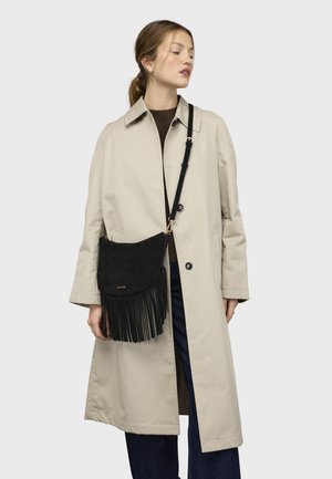 Sac bandoulière en daim noir avec détails en franges et ferrures dorées. Porté avec un manteau beige et un pull foncé, présenté sur un fond neutre.