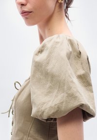 Μπλούζα χακί με φουσκωτά μανίκια και κλείσιμο με κορδόνια μπροστά. Ελαφρύ ύφασμα με ομαλή υφή, διαθέτοντας διακριτικές ραφές και πινελιές με ψιλό σούρωμα.