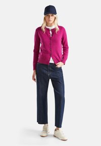 Žena v tmavě modré kšiltovce, magentovém cardigan s knoflíky, bílém tričku, tmavě modrých širokých kalhotách a bílých teniskách stojí proti jednobarevnému pozadí.