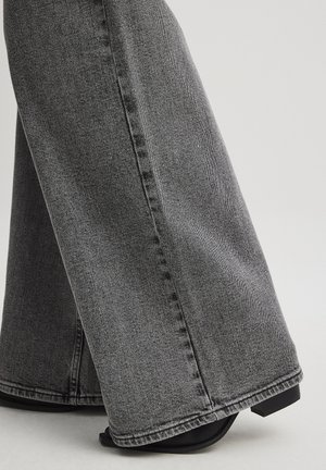 Široke traperice v temno sivi barvi, s gladko teksturo, vidno šivom in čistim robom. Ujemajo se z črnimi sandali na peto.