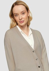 Beige Cardigan aus weichem Stoff mit tiefem V-Ausschnitt, gerippten Kanten und silbernen Knöpfen vorne, kombiniert mit einem weißen Hemd darunter.