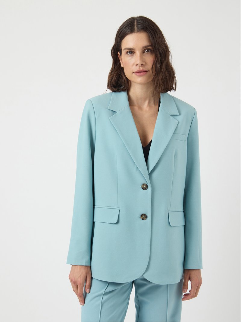 Woman with shoulder-length dark hair wears light blue tailored blazer and matching pants, standing against a plain white background.