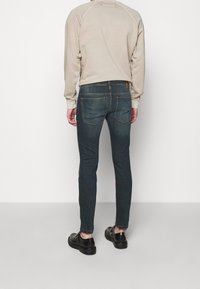 Mörkblå skinny jeans med en klassisk femfickdesign, i kombination med en ljusbeige sweatshirt och svarta skor, sett bakifrån.