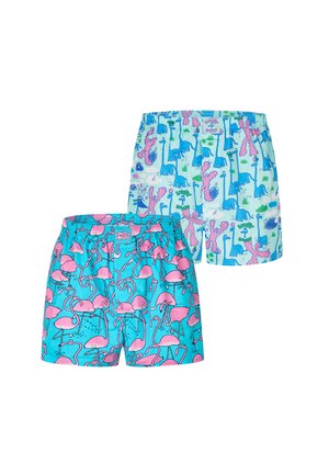 Muskelbepackter Mann mit kurzen Zöpfen, der bunte Shorts mit Dinosaurier-Motiv trägt und vor einem schlichten hellgrauen Hintergrund steht.