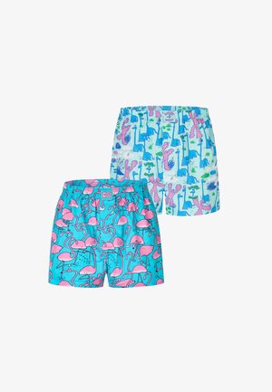 Muskelbepackter Mann mit kurzen Zöpfen, der bunte Shorts mit Dinosaurier-Motiv trägt und vor einem schlichten hellgrauen Hintergrund steht.
