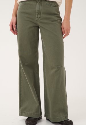 Personne portant un pantalon vert olive à jambes larges et des chaussures noires, debout avec les mains détendues sur les côtés devant un fond clair et uni.