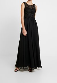 Svart klänning med en spetsliv och pärlbesatt midja. Flödar ut i en plisserad kjol. Ärmlös design med rund halsringning. Slät tygtextur.