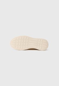 Sole of a beige rubber shoe with grid and linear tread patterns, featuring a small embossed logo near the arch area.