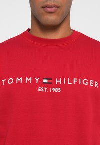Felpa rossa in cotone con ricamo bianco "TOMMY HILFIGER EST. 1985" e un accento di logo in bianco e nero. Collo a giro classico.
