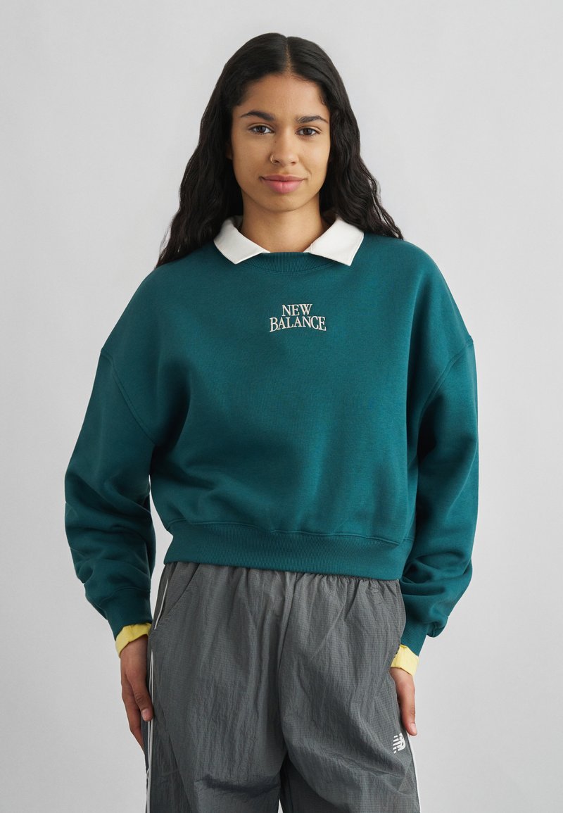 Γυναίκα με μακριά σκούρα μαλλιά, που φοράει μπλε-πράσινο φούτερ New Balance πάνω από λευκό πουκάμισο με γιακά και γκρι αθλητικό παντελόνι σε ανοιχτό γκρι φόντο.