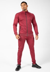 Rode sportieve tracksuit met een ritsjack en aansluitende broek. Gemaakt van textuurstof, met zwarte accenten en twee zijzakken.