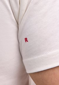 Weißes Kurzarmhemd mit weicher Textur. Verfügt über ein kleines rotes "R", das auf dem Ärmel gestickt ist. Die Nahtdetails sind sauber genäht.