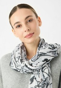 Écharpe légère et blanche avec un motif floral noir, texture douce, drapée autour du cou, complétant un pull gris.