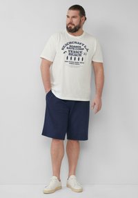Witte katoenen t-shirt met blauwe grafische tekst, gecombineerd met marineblauwe shorts tot de knie. Model draagt witte sneakers en staat tegen een neutrale achtergrond.