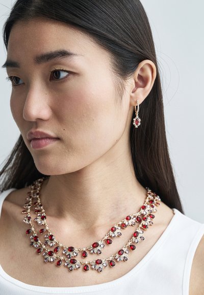Raudona ir skaidrių brangakmenių kaklo papuošalas su sluoksniuota struktūra, su auksinės spalvos grandinėle. Suderinti raudoni brangakmenių auskarai su dekoratyviniais akcentais.