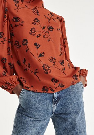 Lang mouwen blouse in roestkleur met zwart bloemmotief, met opgeblazen mouwen en elastische manchetten, gecombineerd met lichtblauwe spijkerbroek. Handen in de zakken.