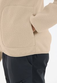 Beige fleecejacka med en texturerad yta och kontrasterande detaljer, bära över mörka byxor med sidoficka och slät tyg.
