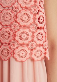Korallenfarbenes Spitzentop mit floralem Stickerei und gewellten Kanten, über einem weichen, rosa, strukturierten Stoff geschichtet. Detailliertes Design mit kreisförmigen Motiven.