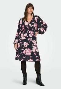 Žena u crnoj haljini do koljena s ružičastim cvjetnim uzorkom, dugim napuhanim rukavima, crnim tajicama i crnim čizmama do gležnja, stoji samouvjereno.