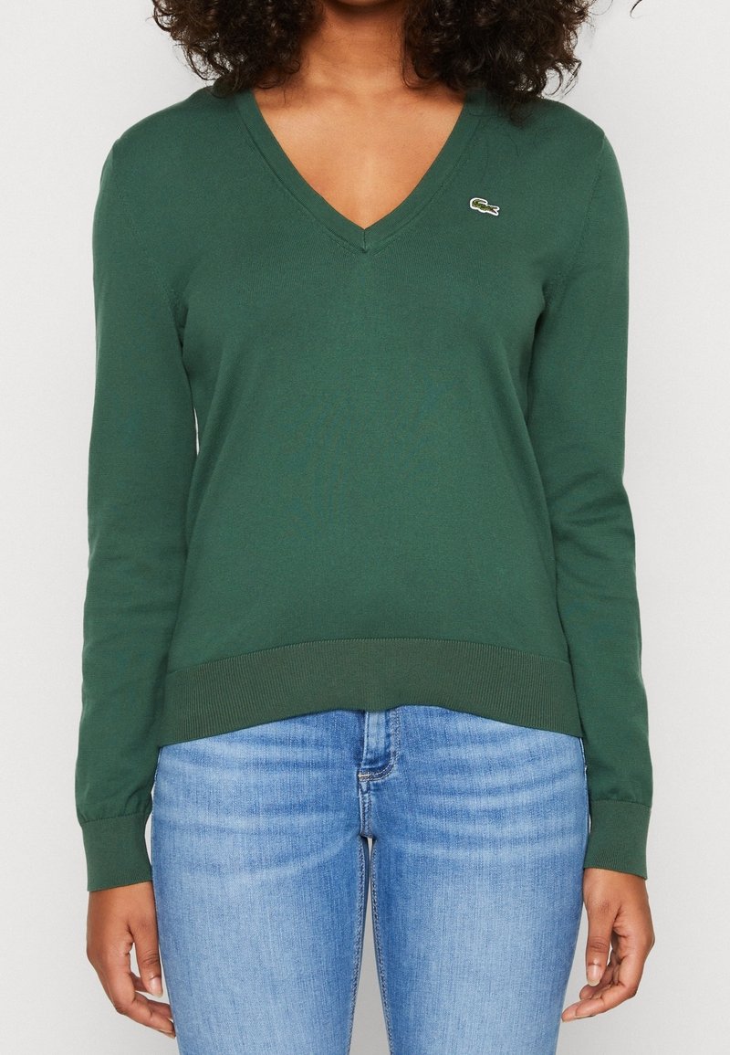 Grön v-ringad tröja, stickad tyg, figurnära långa ärmar, ribbade manschetter och fåll, med en liten broderad logotyp på bröstet.