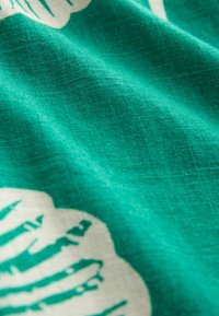 Textil szövet texturált, szőtt felülettel élénk türkizkék színben, fehér levélmintákkal díszítve. A mintázat merész és élesen kirajzolódó.