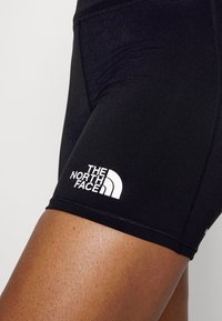 Černé sportovní šortky vyrobené z elastického materiálu, s bílým logem na boku a hladkým povrchem se zřetelnými švy.