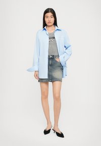 Camicia oversized azzurro chiaro con bottoni, canotta a righe grigie, gonna di jeans, mule neri senza tacco. Tessuto morbido, look a strati informale.