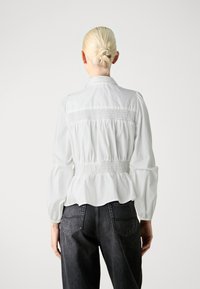 Weiße Bluse mit langen Ärmeln, gerafftem Bund und Raffung, mit Kragen und einem leicht ausgestellten Saum. Getragen mit dunkelgrauen Jeans.