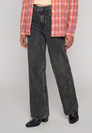 Personne portant un jean noir à jambes larges, des bottines noires et une chemise à carreaux rouge avec les manches retroussées.