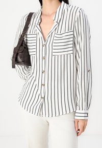 Femme portant une chemise blanche boutonnée avec des rayures noires verticales et deux poches poitrine rayées horizontales, portant un petit sac à bandoulière marron foncé.