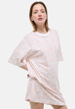 Femme portant une robe ample à manches courtes avec un motif floral rose pâle, debout une main sur la hanche devant un fond blanc.