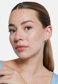 Goldene Creolen mit einem strukturierten Design, getragen von einer Person mit glatter Haut und einem hellblauen Oberteil. Die Haare sind nach hinten gegelt.