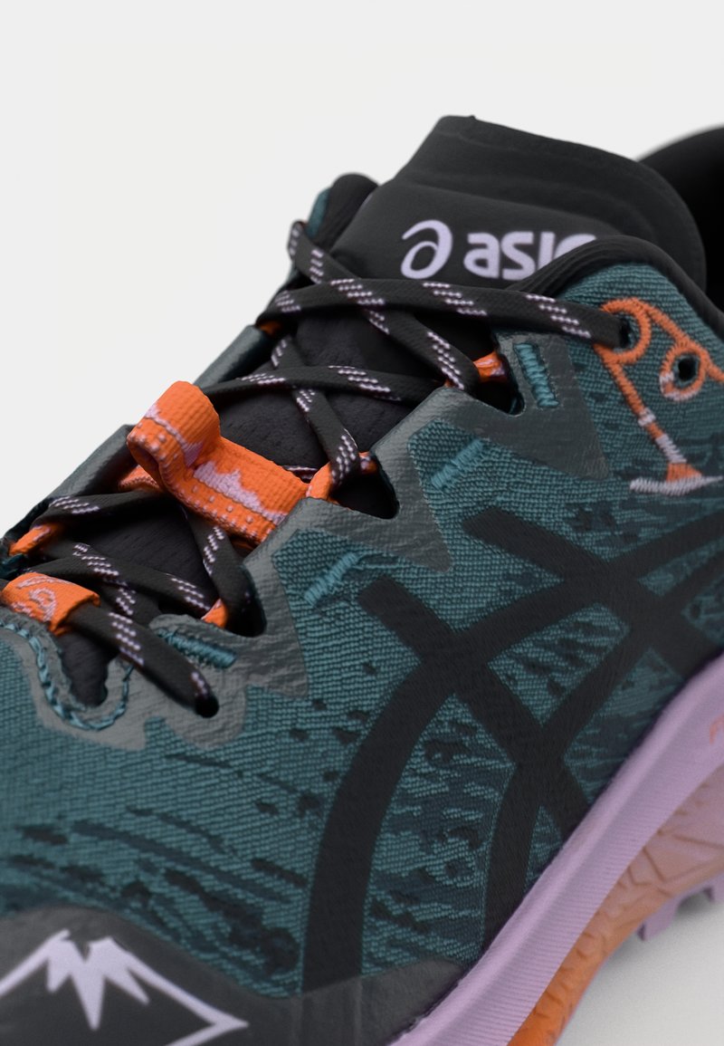 Buty do biegania po szlakach mają teksturowaną, turkusową cholewkę z czarnymi akcentami i pomarańczowymi detalami. Cechą wyróżniającą jest wzorzysty design i fioletowa podeszwa.