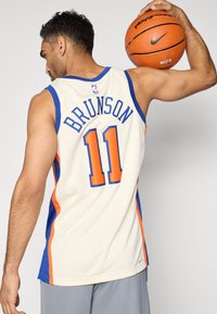 Basketball-Trikot in Creme mit blauen und orangefarbenen Akzenten, auf der Rückseite mit "BRUNSON" und der Nummer "11" bedruckt. Spieler hält einen Basketball.